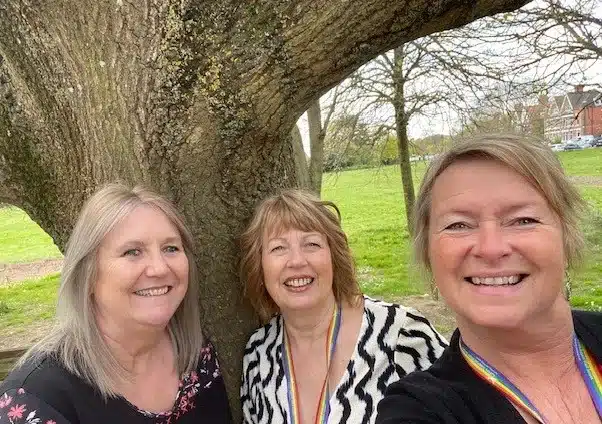 Three carer coaches standing by large tree smiling wearing rainbow lanyards
